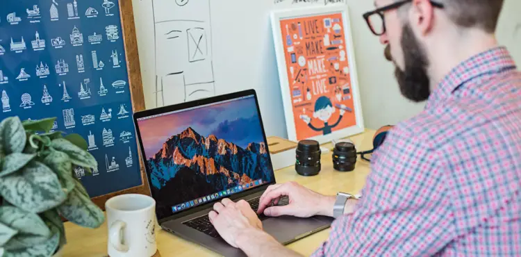 A creative professional or developer working on a laptop at a brightly lit desk with design artwork and camera lenses in the background.