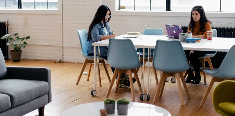 Two marketing professionals working on laptops at a collaborative workspace table in a bright, modern studio office.