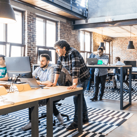 Two male colleagues collaborating at a desk on a computer inside a bustling, modern industrial-style co-working office with brick walls.