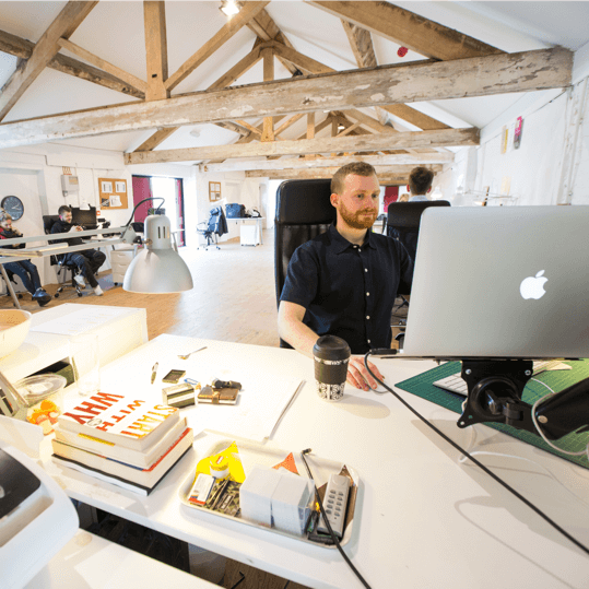 A male professional working on a laptop at a desk in a spacious, modern open-plan office with rustic exposed wooden ceiling beams.