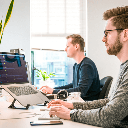 A male software engineer coding on a laptop laptop stand at an office desk with a colleague working in the background.