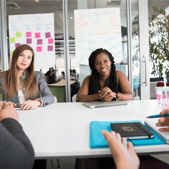 Two female business professionals listening in a meeting with a MoSCoW method prioritisation chart and a customer journey map on the glass wall behind them