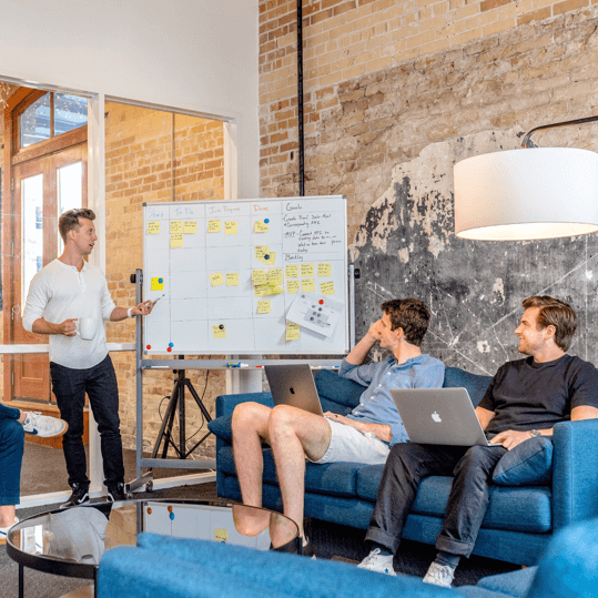 A presenter leading an agile sprint planning session with a whiteboard Kanban board while team members listen on a blue sofa.