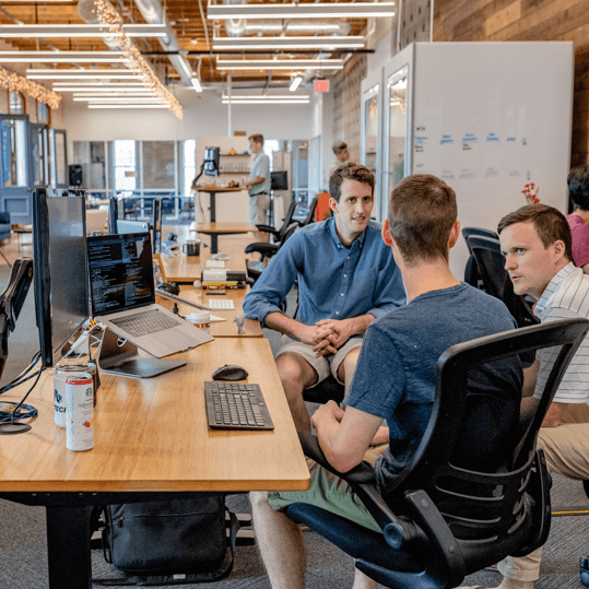 A group of male tech professionals collaborating and discussing a software development project at a long wooden desk in an open-plan office.