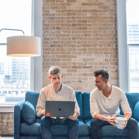 Two male colleagues collaborating on a laptop while sitting on a blue sofa in a modern office with a brick accent wall.