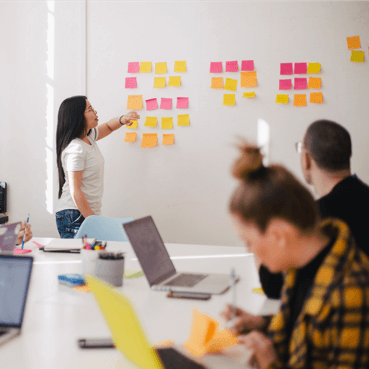 Female presenter leading an agile design thinking workshop with team members looking at colourful sticky notes on a wall.