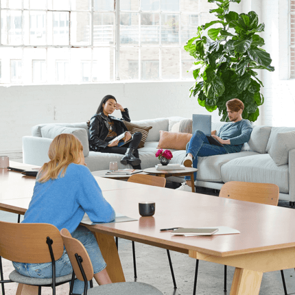 Professionals working and relaxing in a bright, modern open-plan coworking space with a large sectional sofa and a wooden conference table.