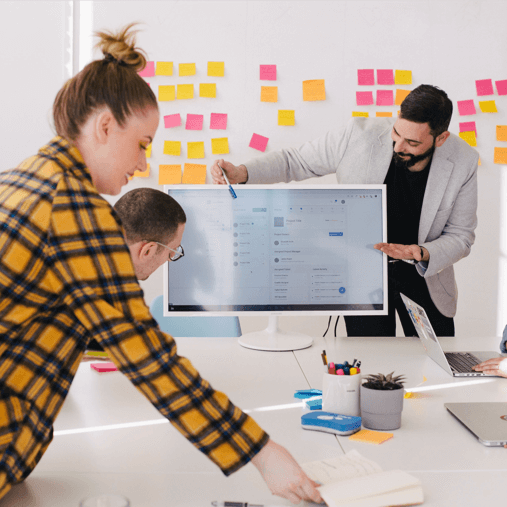 A male project manager presenting a digital software dashboard on a monitor to his team during an agile sprint planning meeting.